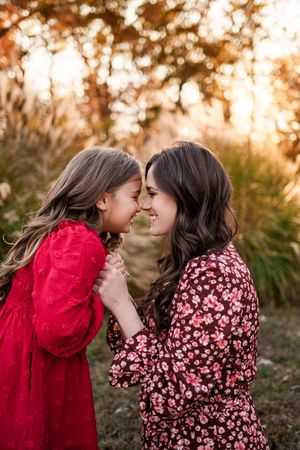 Mother and daughter in red and floral dresses touching noses and smiling in fall park.