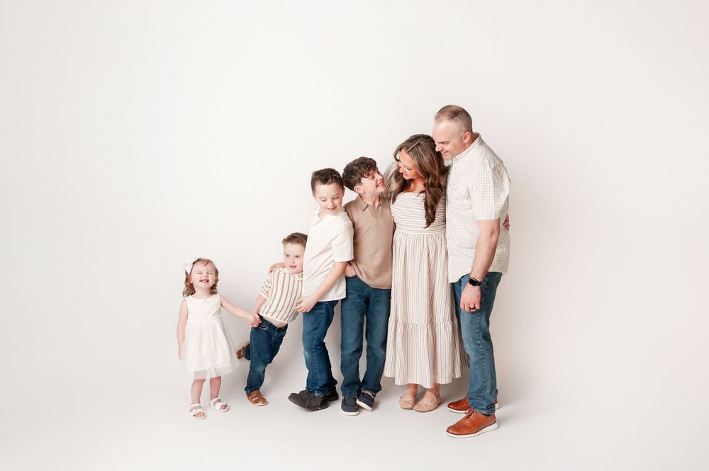 A family of six poses playfully in soft neutral tones against a clean white studio backdrop.