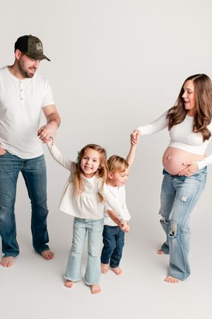 Pregnant mother and father hold hands with two young children in a bright white studio family portrait.