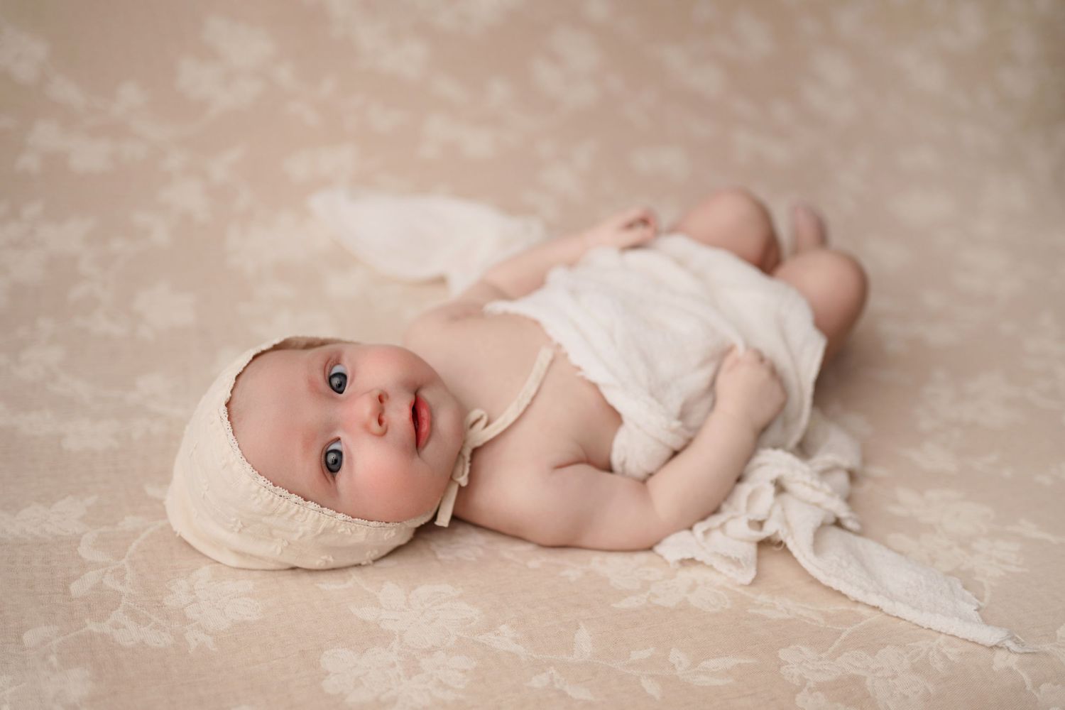Smiling baby girl in white lace dress and bow headband doing tummy time on a white quilted bed.
