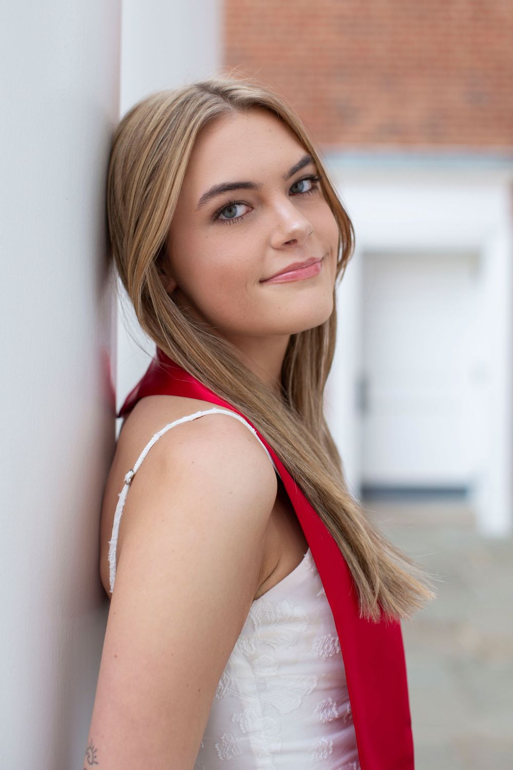 Blonde young woman in white dress with red graduation stole leaning against white pillar outdoors.