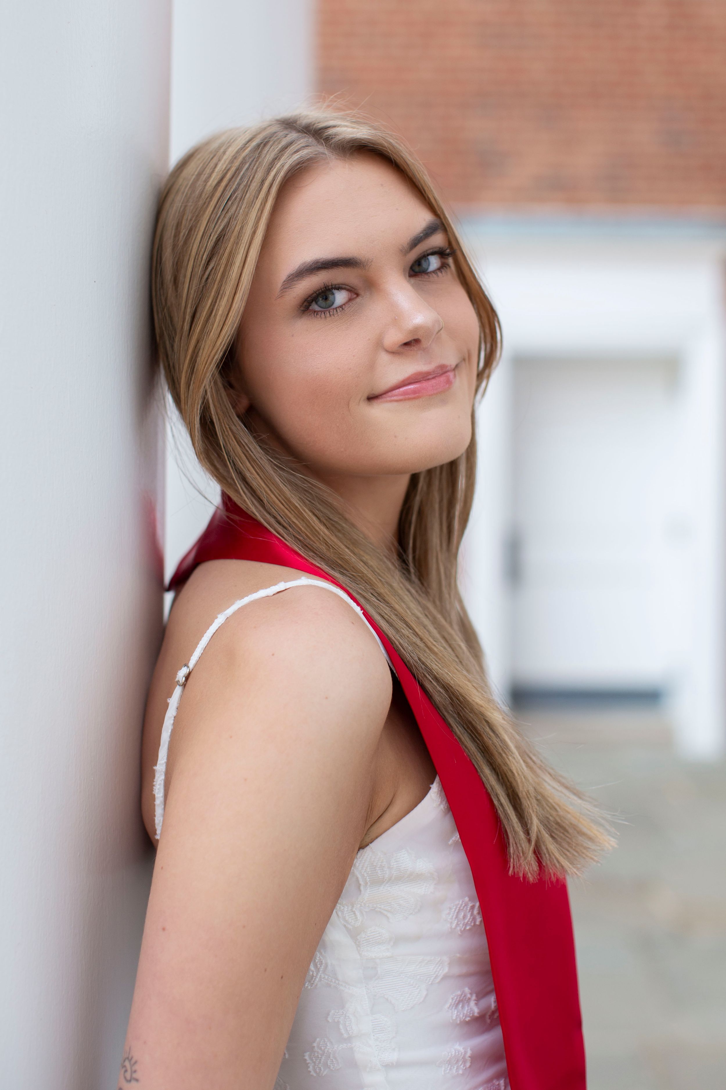 Blonde young woman in white dress with red graduation stole leaning against white pillar outdoors.