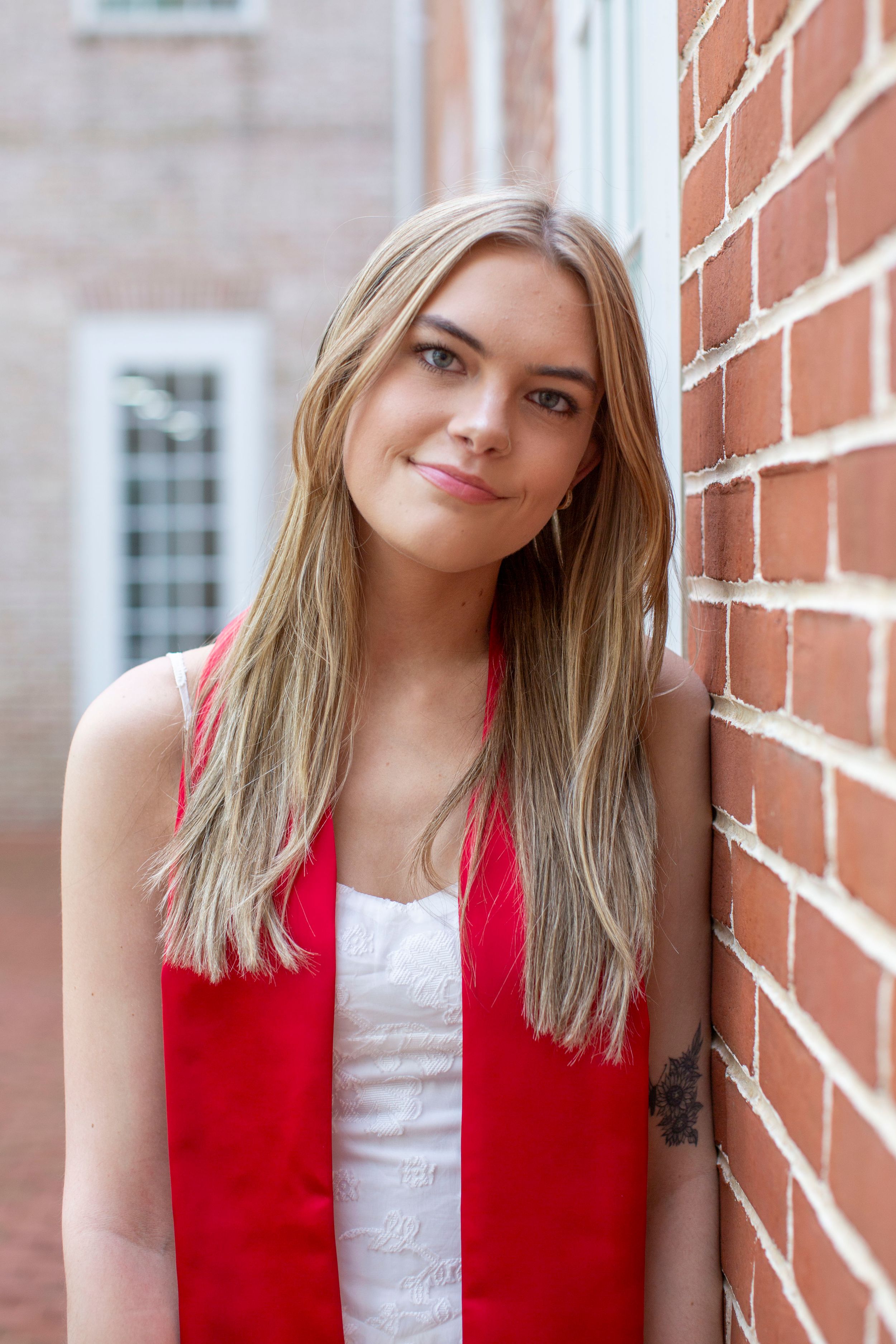 Smiling blonde graduate in white dress and red stole leaning against brick wall outdoors.