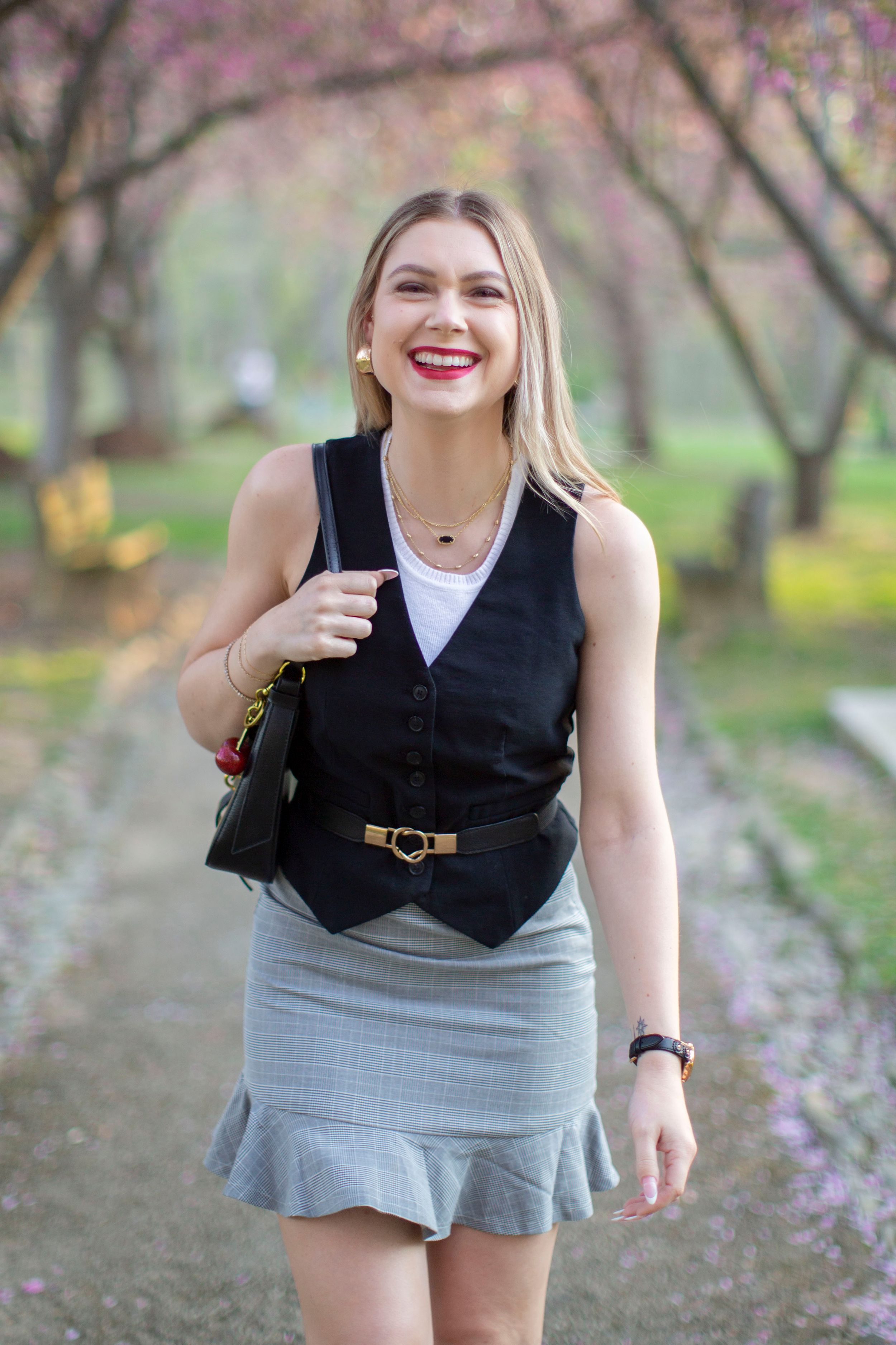 Laughing blonde woman in black vest and gray skirt walking through pink cherry blossom park.