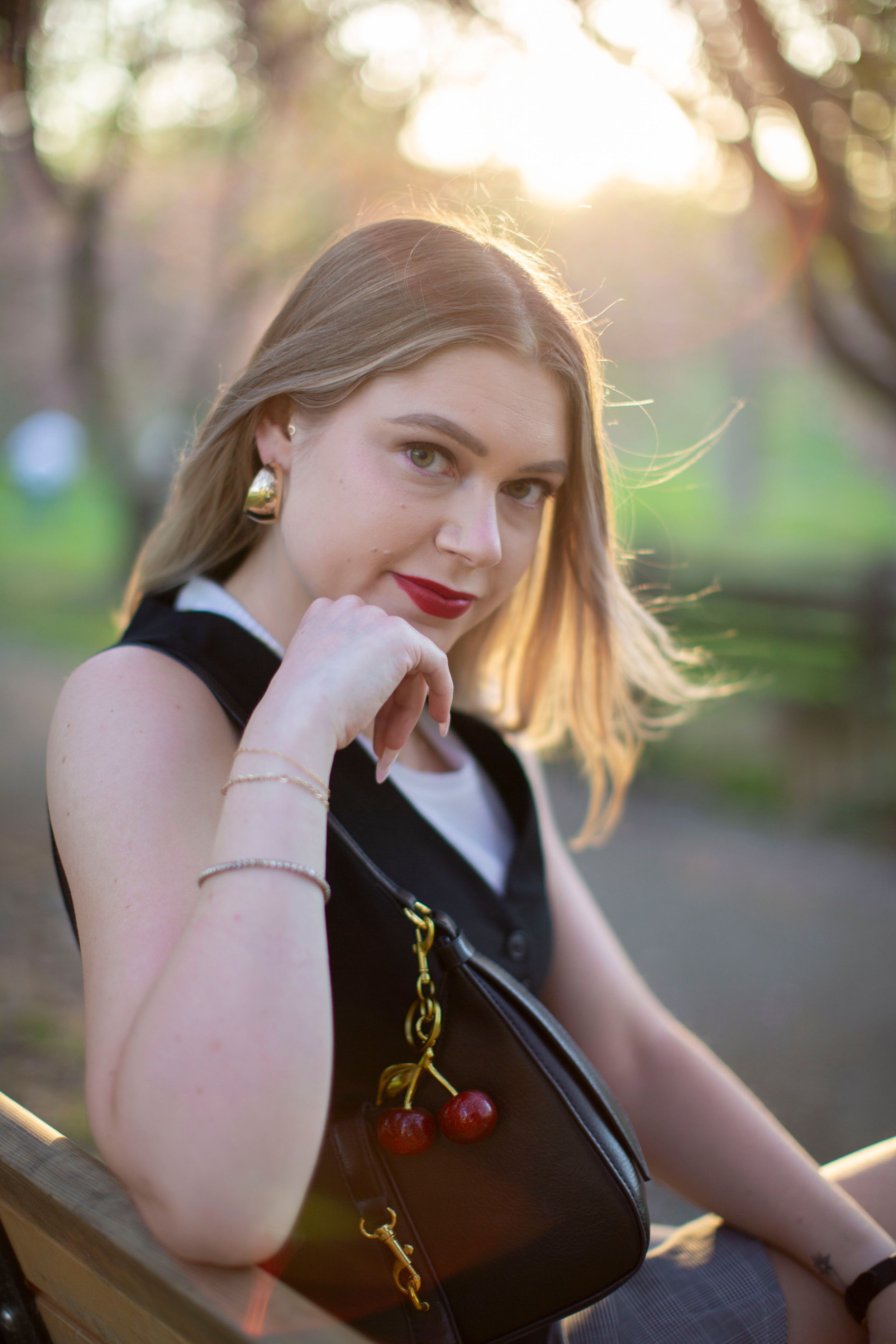 Blonde woman in black vest with cherry bag charm sitting on bench at golden hour.