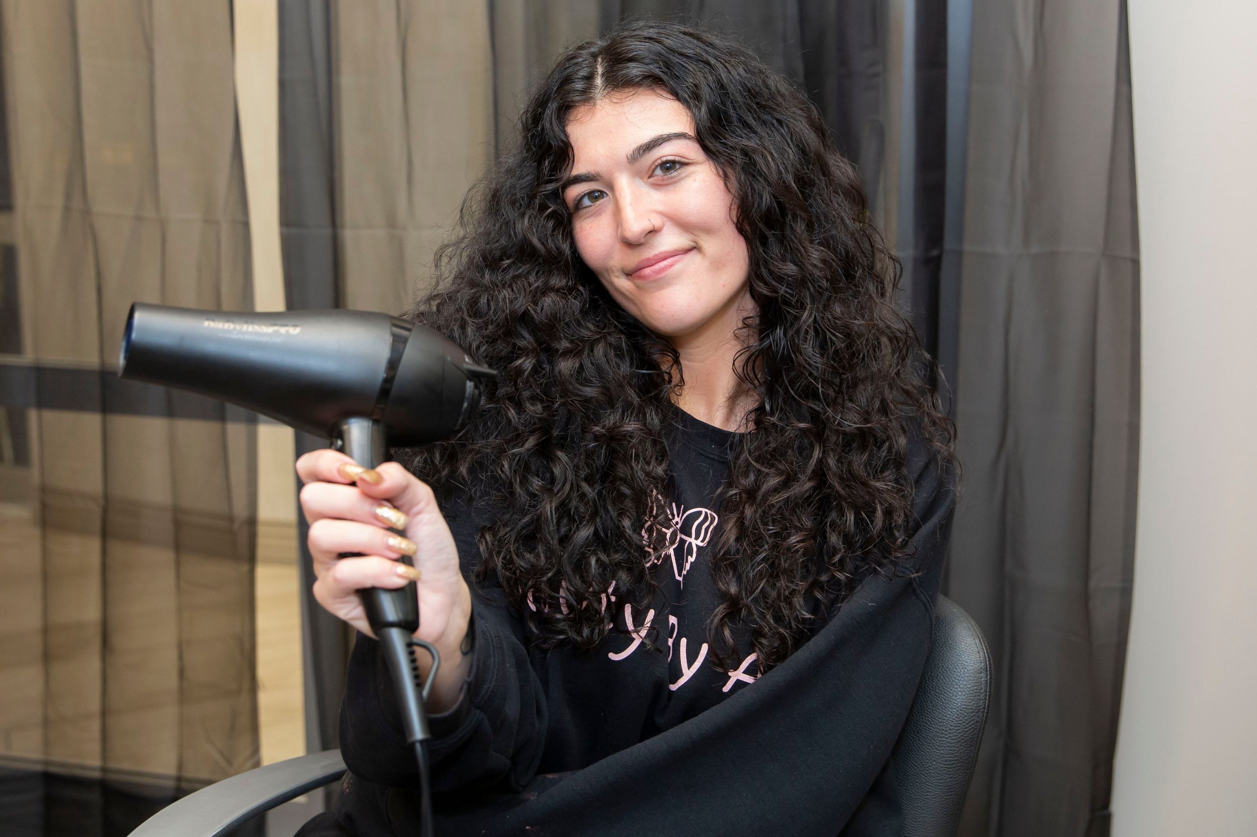 Young woman with curly dark hair holding a hair dryer at a salon.