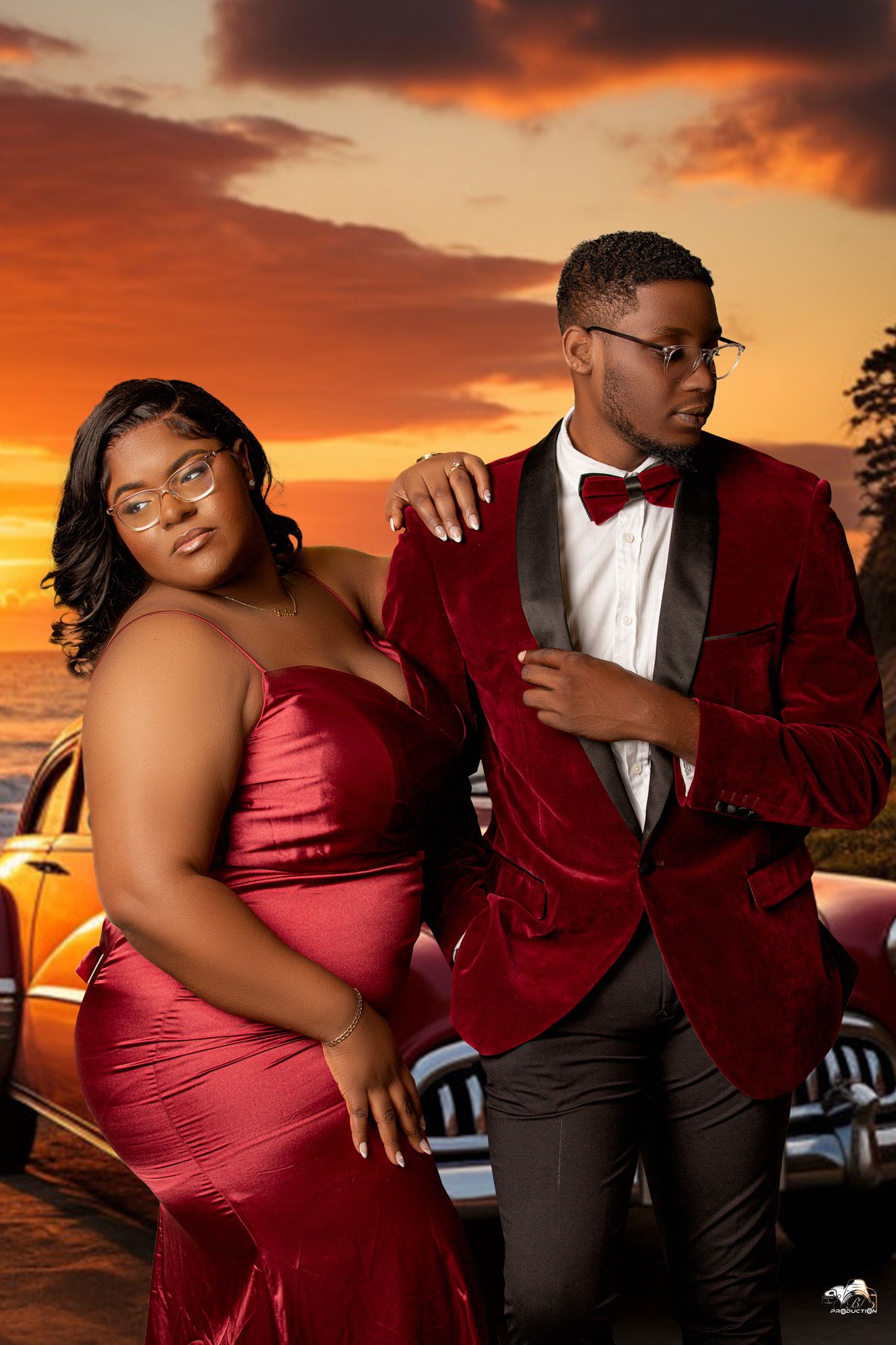 A couple in formal attire poses against a dramatic sunset sky, with her in red and him in a burgundy tuxedo jacket.