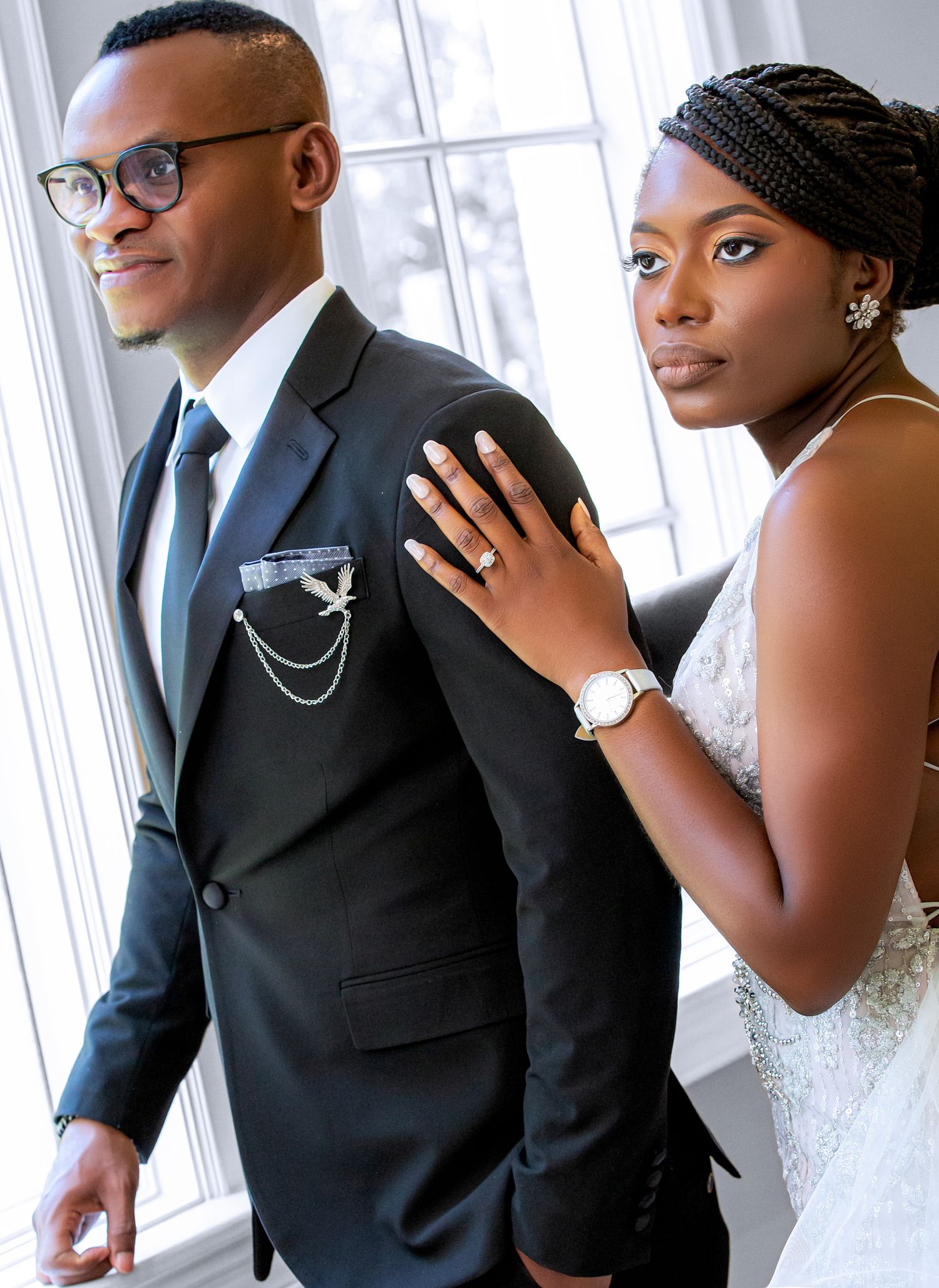 An elegant couple poses by a window in wedding attire featuring a black suit and white gown.