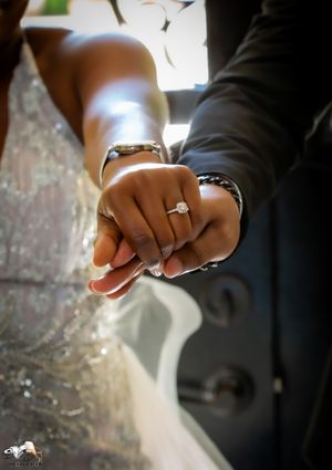 Close-up shot of hands displaying wedding rings against a white dress and dark suit.