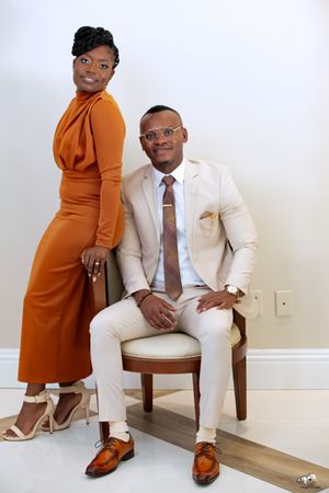 A stylish couple poses in an orange jumpsuit and cream suit against a white studio backdrop.