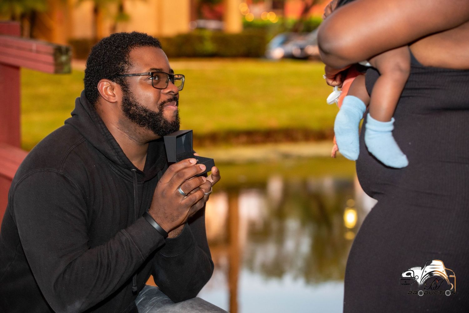 A romantic proposal scene by a lake with tiny baby shoes held in the background.