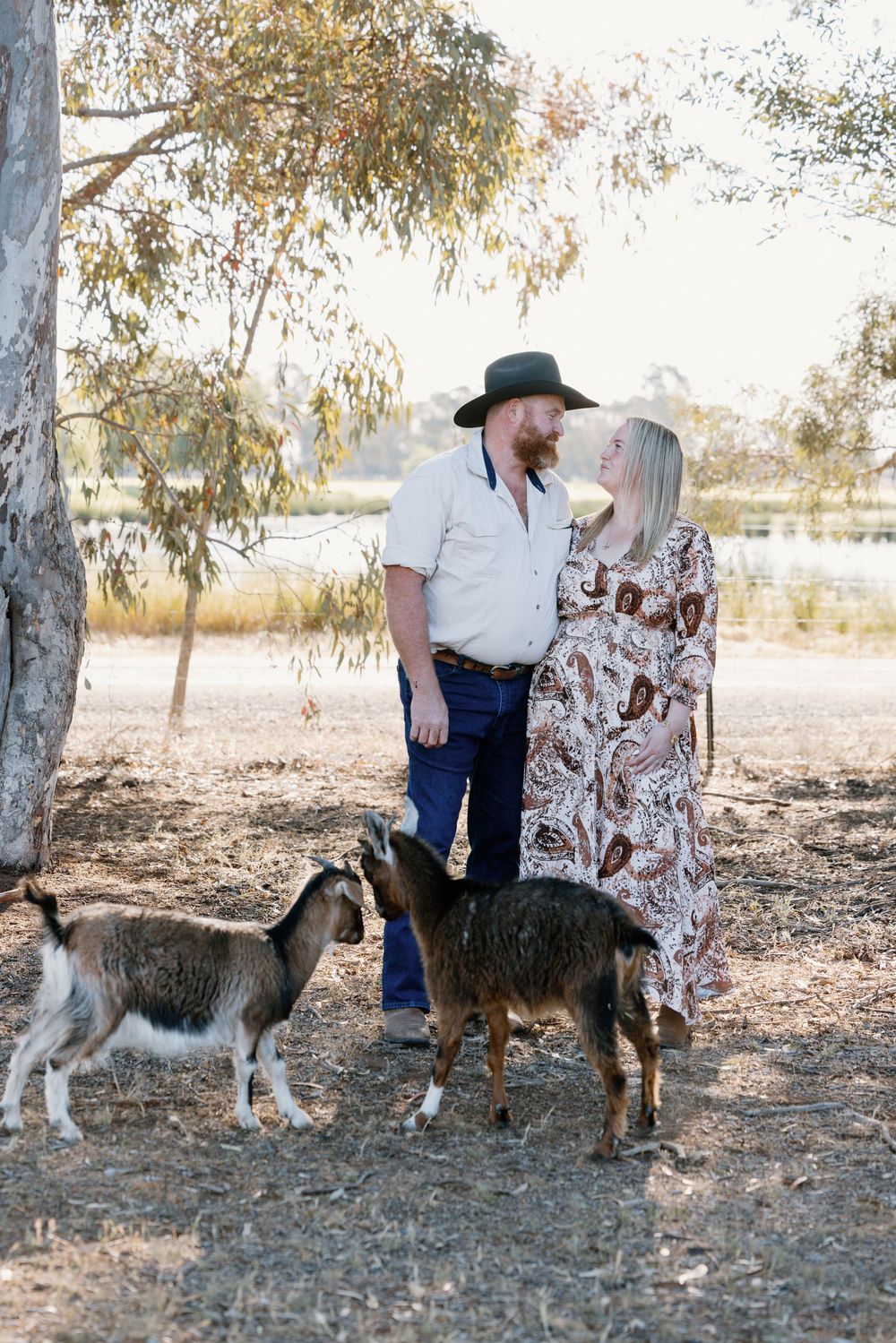 A couple stands together under eucalyptus trees in a rural setting with two goats beside them.