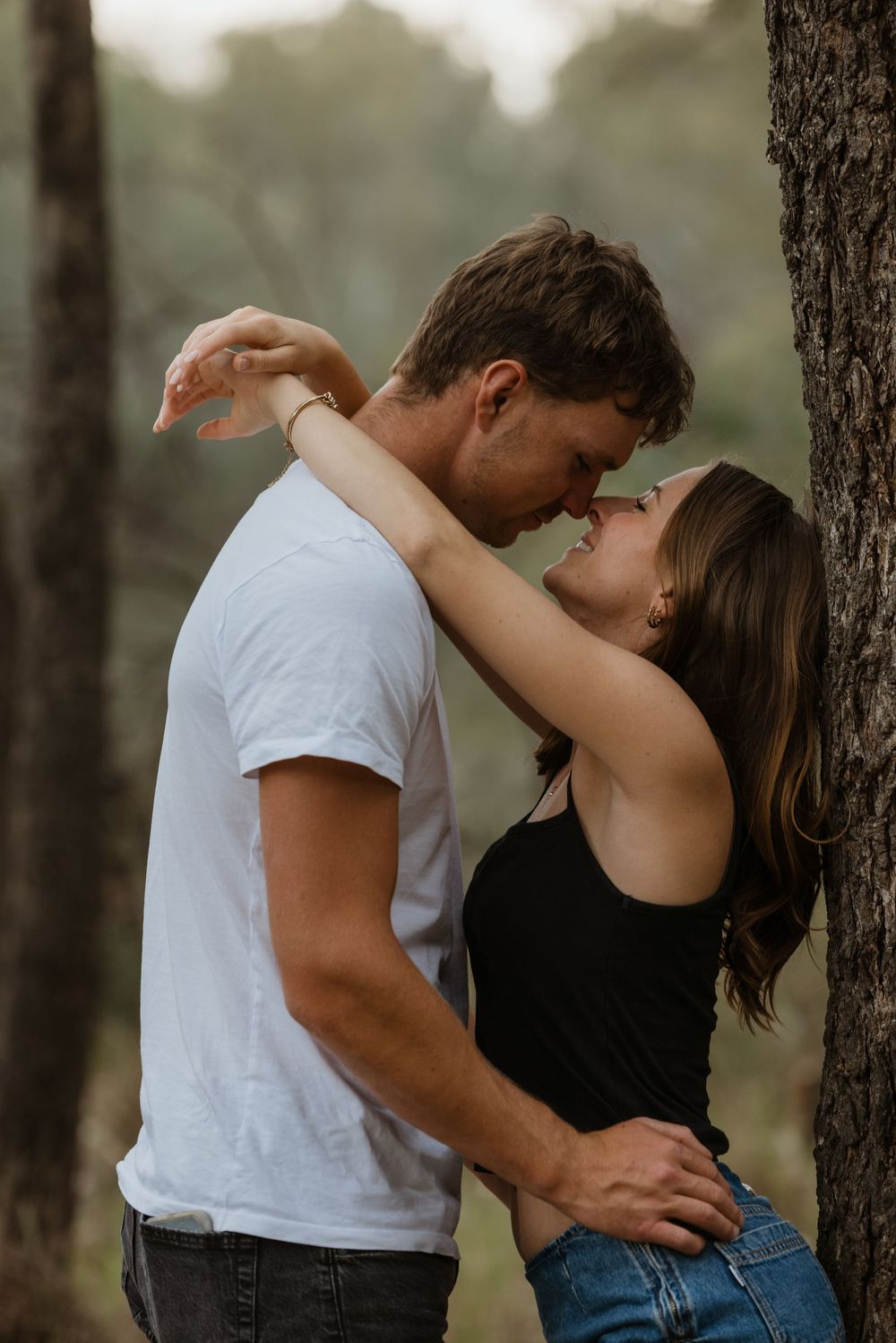 Romantic couple embraces passionately against tree trunks in a misty forest setting.