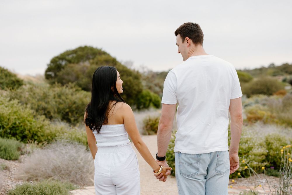 A couple in white outfits holding hands while walking on a nature path surrounded by green foliage.