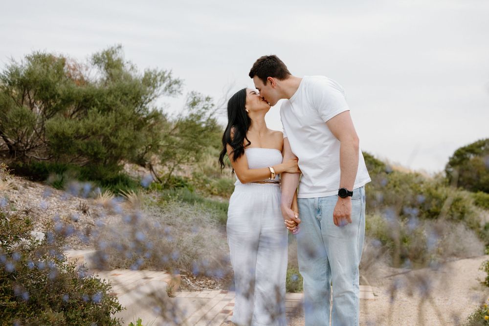 A couple embraces while standing on a rocky coastal trail during sunset with ocean views in the background.