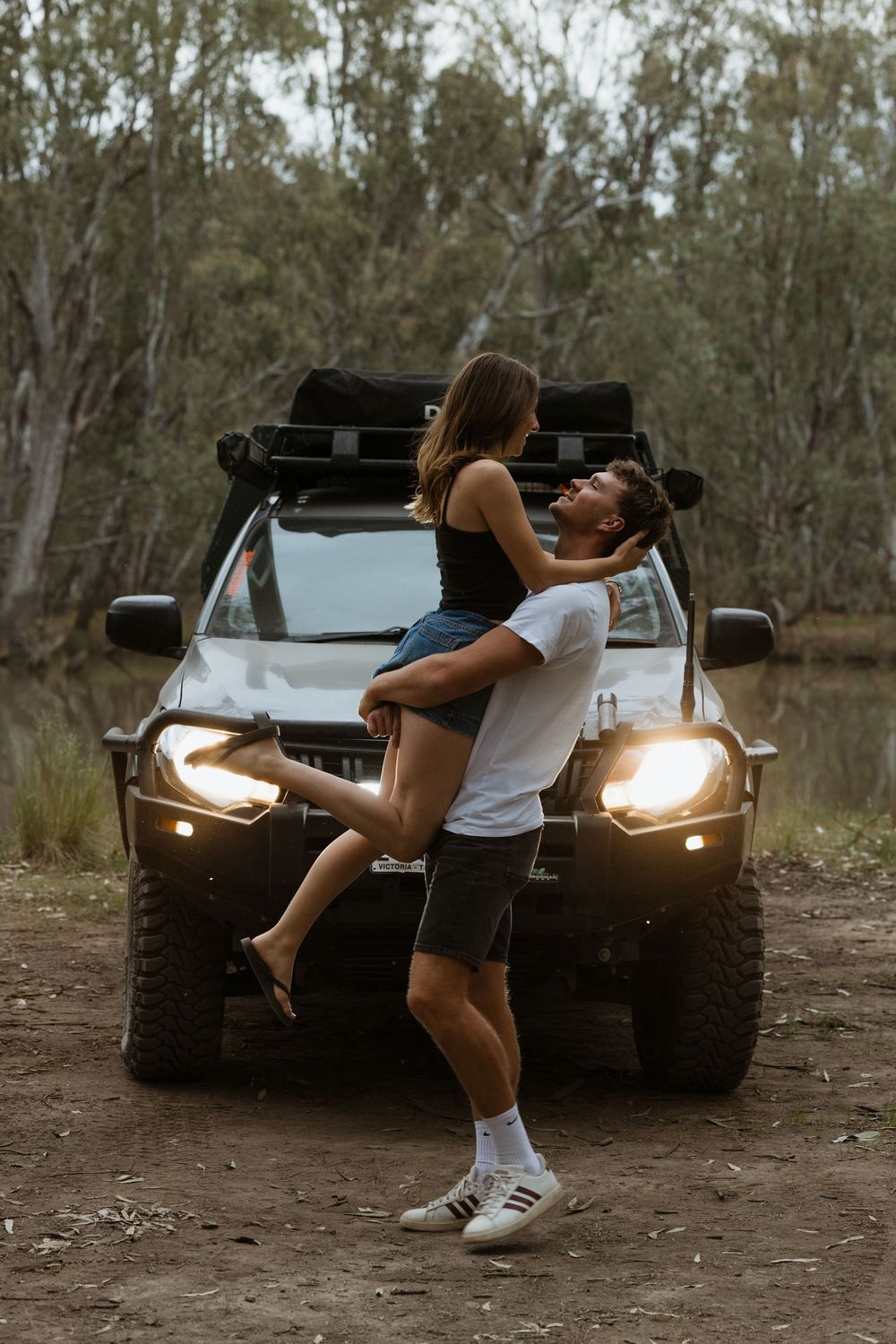 A woman in white tank top and shorts poses against a black Jeep with headlights on at dusk in a wooded area.