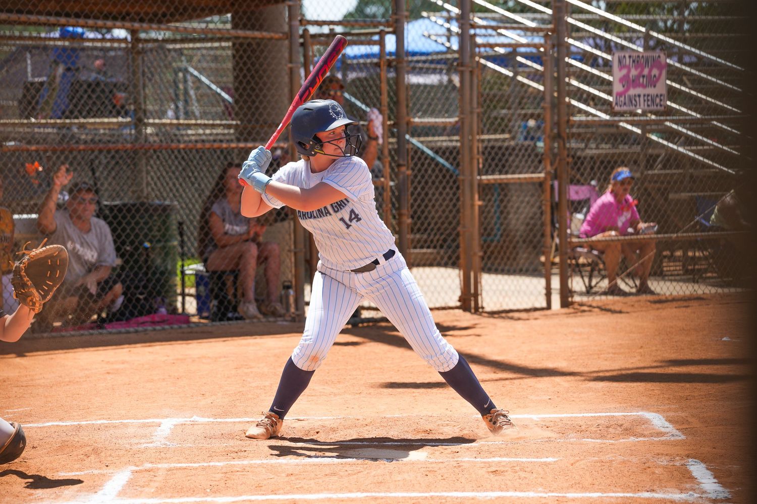 Baseball player in batting stance at home plate with spectators visible through fence.