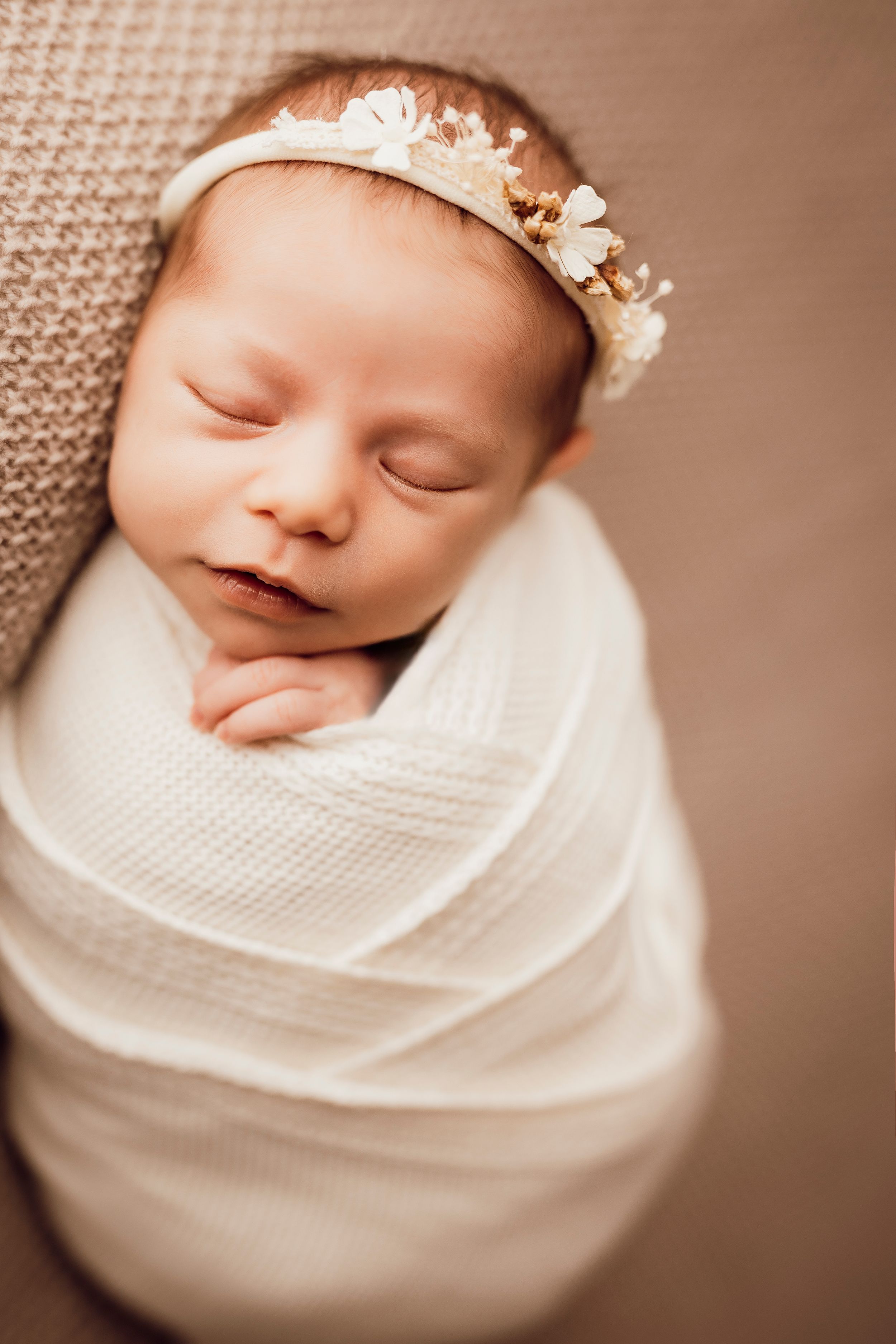 Newborn girl with a white floral headband sleeps swaddled in a cream wrap against a soft neutral knit blanket.