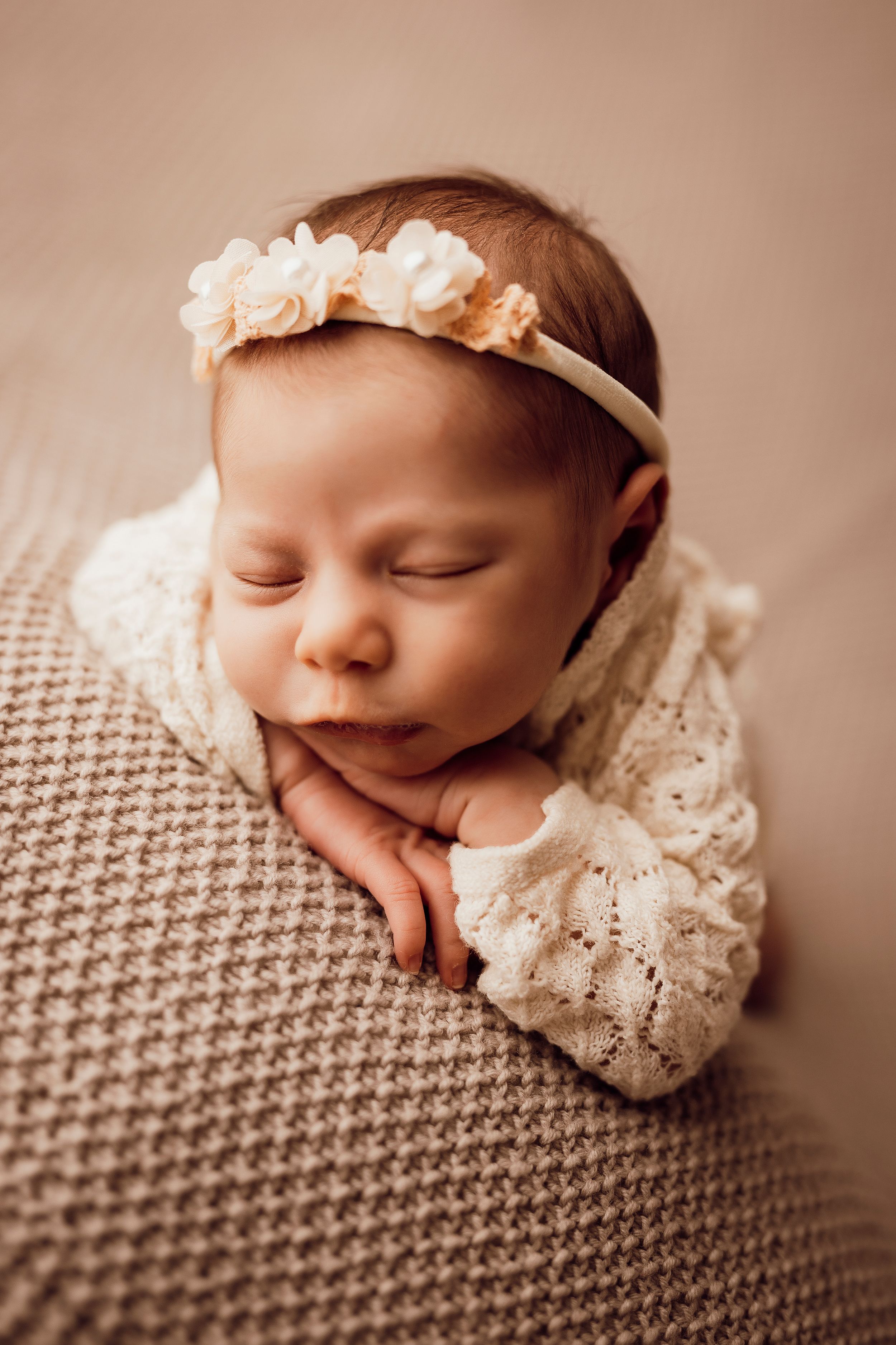 Sleeping newborn in a lace outfit and peach flower headband rests her chin on her hands on a textured beige knit blanket.
