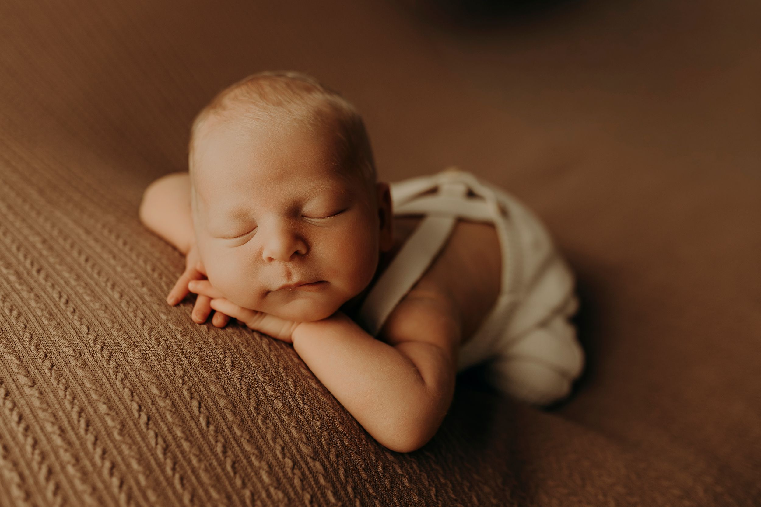 Sleeping newborn baby rests his head on folded arms atop a textured brown blanket in a peaceful newborn portrait.