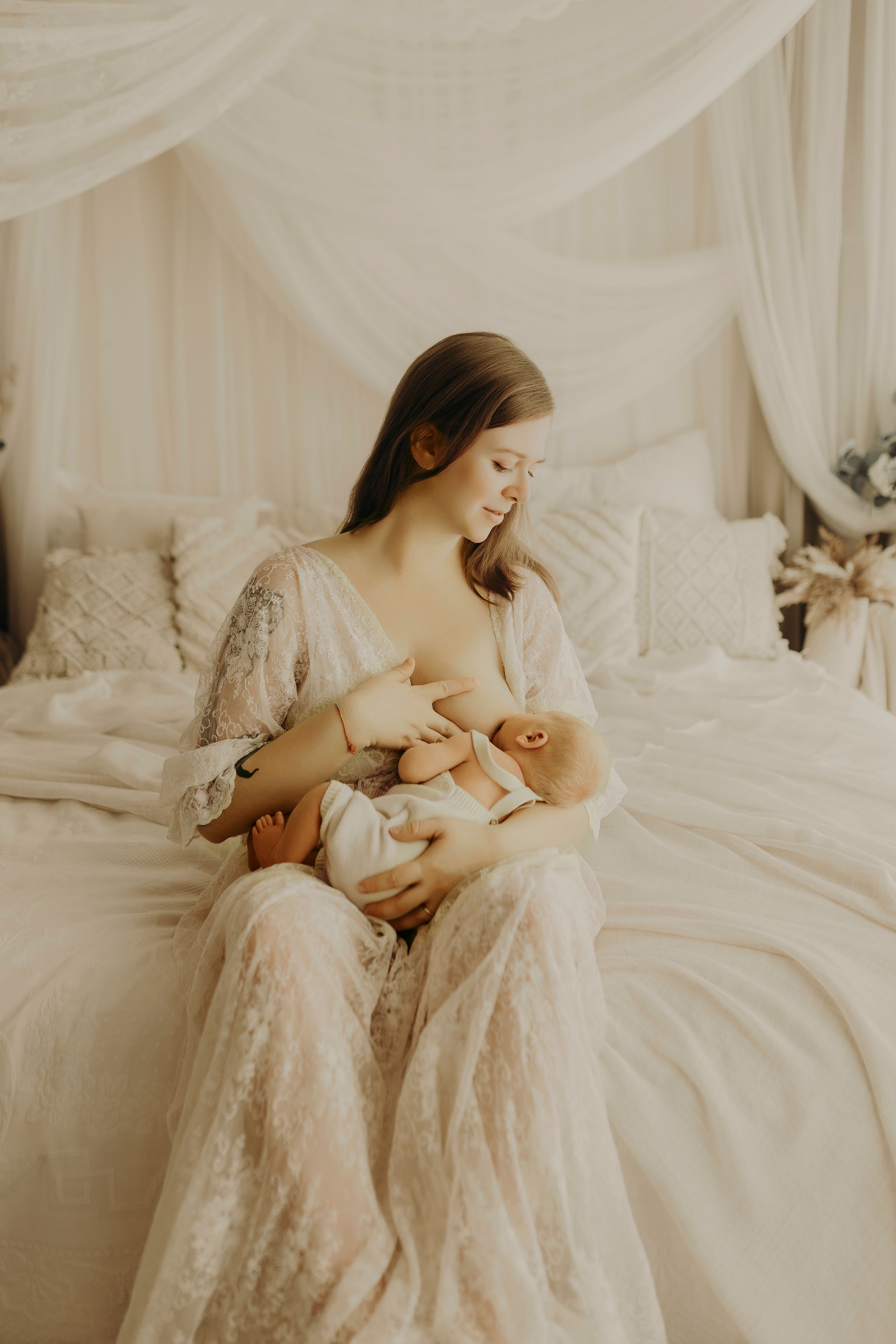 Mother in a flowing lace gown breastfeeds her newborn on a white bed draped with sheer canopy fabric in a serene lifestyle photoshoot.