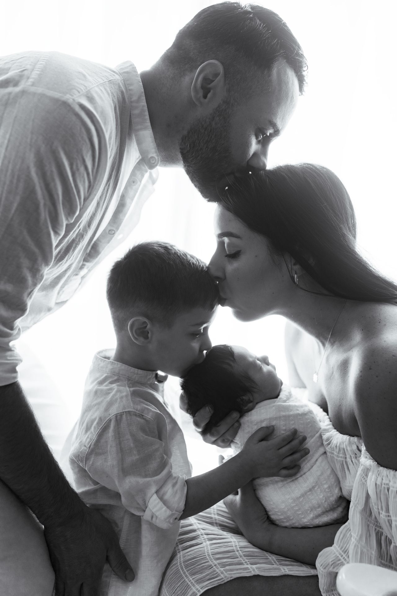 Black and white newborn family portrait of parents and a young boy kissing and gently touching a swaddled baby.