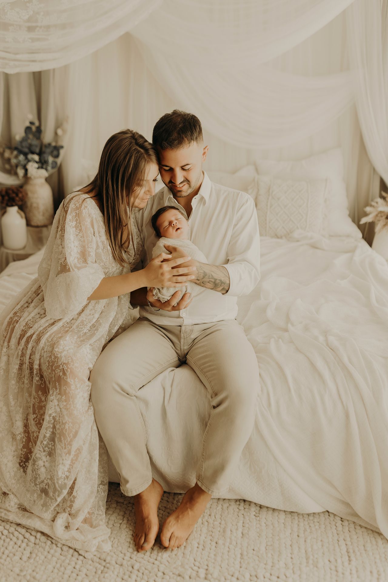 New parents in cream outfits sit on a white canopy bed gazing lovingly at their swaddled newborn in a cozy family photo.