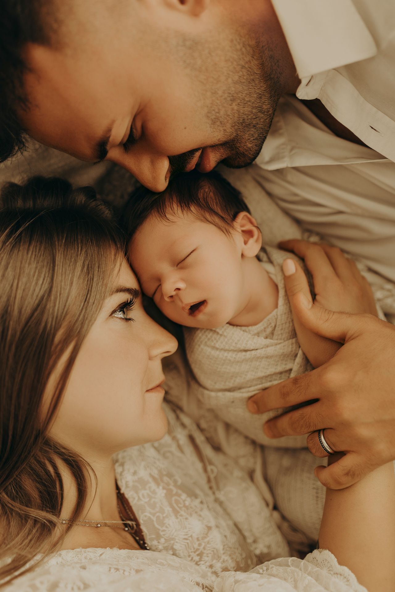 Close-up of a sleeping newborn nestled between mom and dad as the father gently kisses the baby's head in an intimate family portrait.