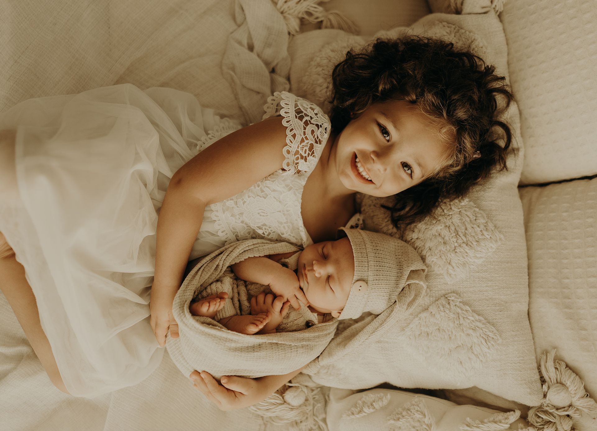 Smiling curly-haired girl in a white dress lies on a soft bed cuddling a sleeping newborn baby in a cream wrap.