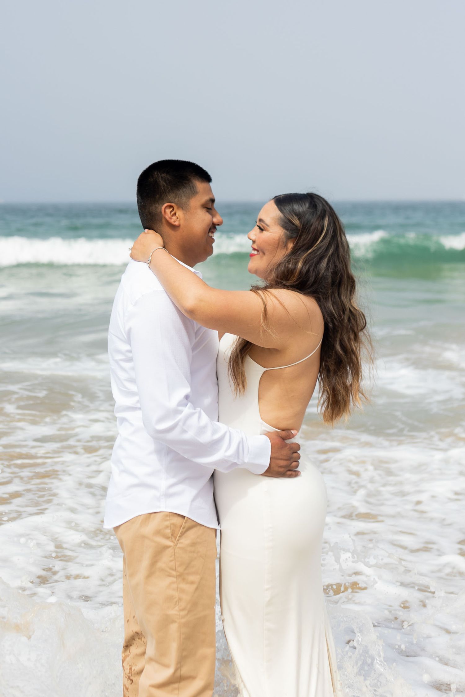 A couple embraces lovingly on a beach with crashing waves in the background.
