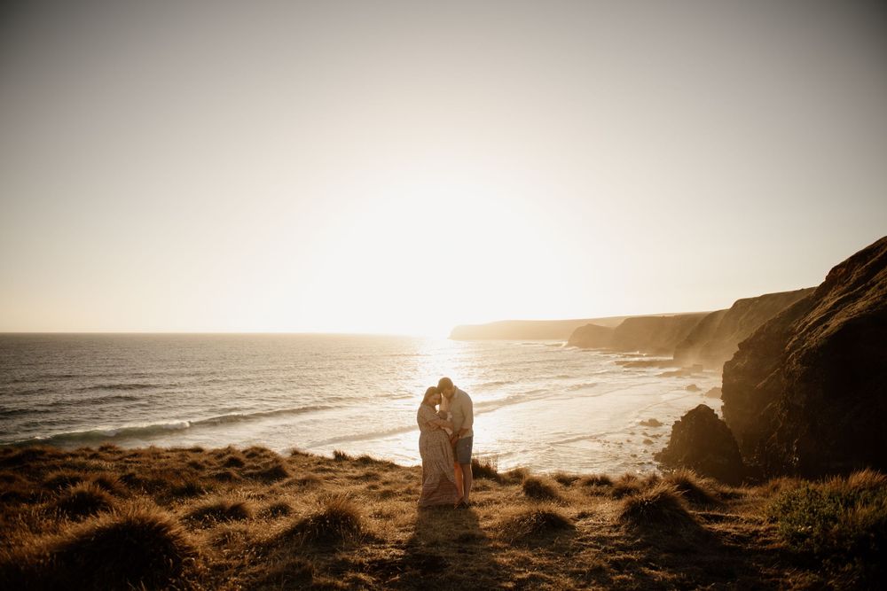 A silhouetted couple shares a romantic moment at sunset on a dramatic coastal cliff overlooking the ocean.