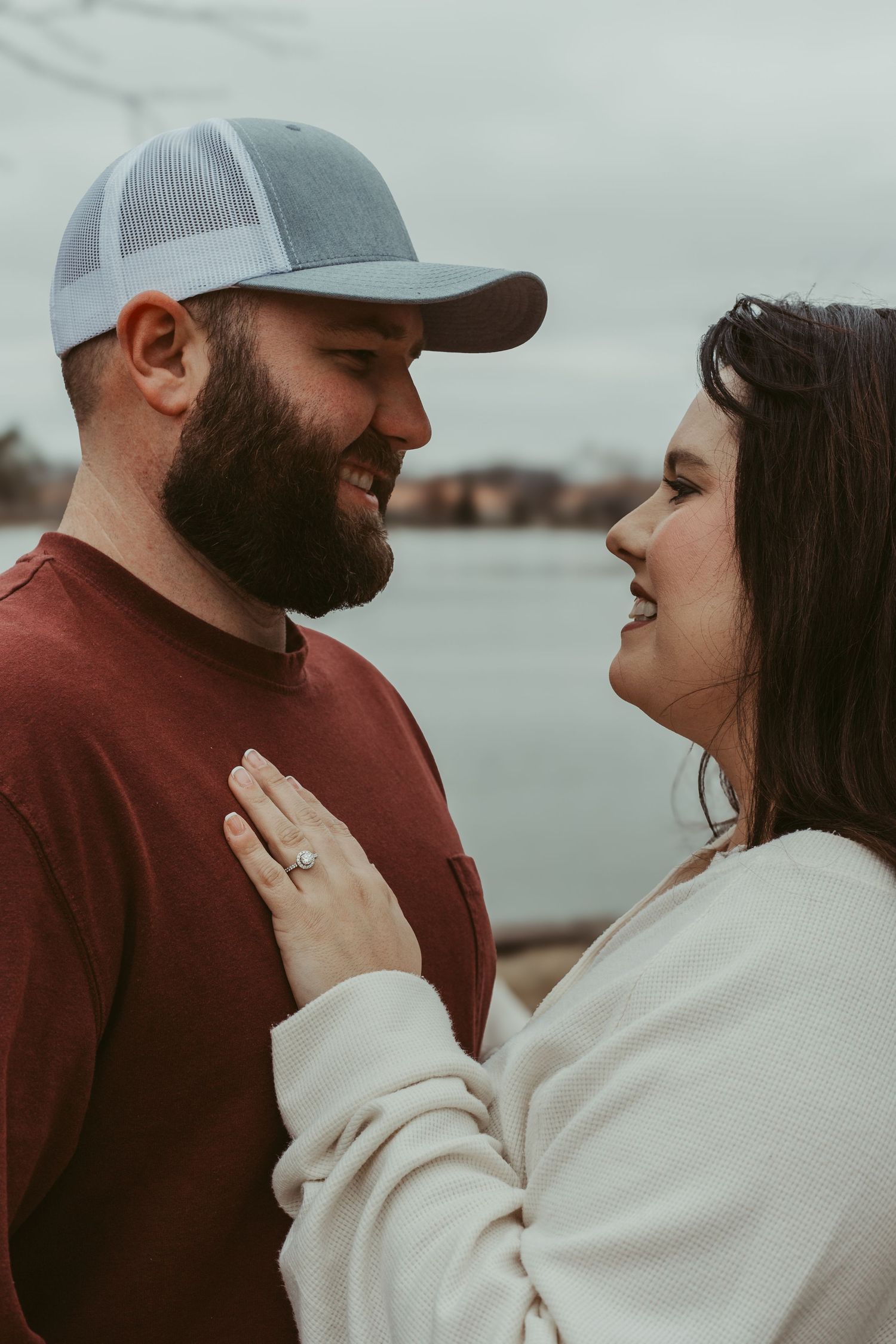 A couple shares an intimate moment together by a lake, with the woman showing off her engagement ring.