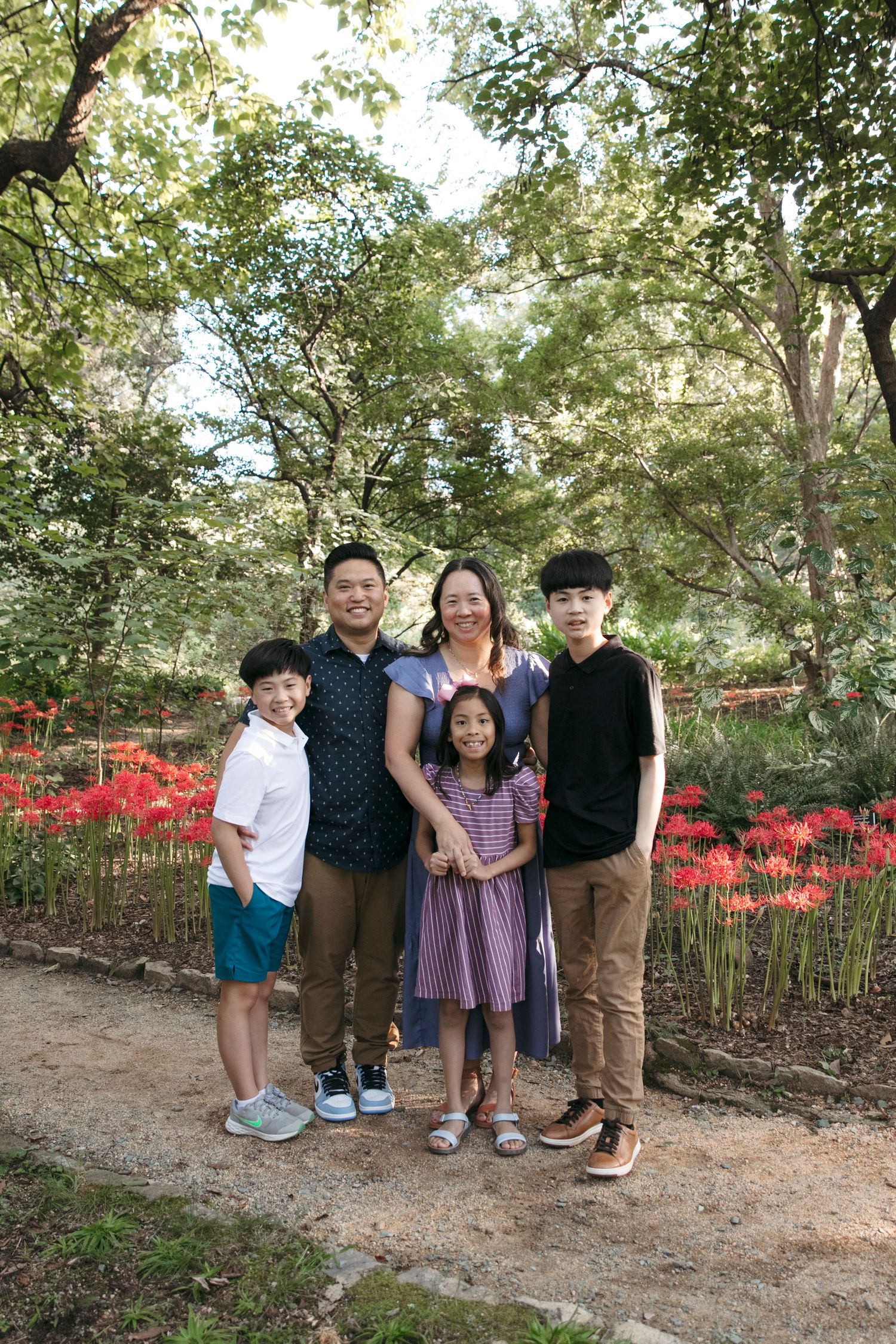 A family stands together on a path surrounded by trees and vibrant red flowers.