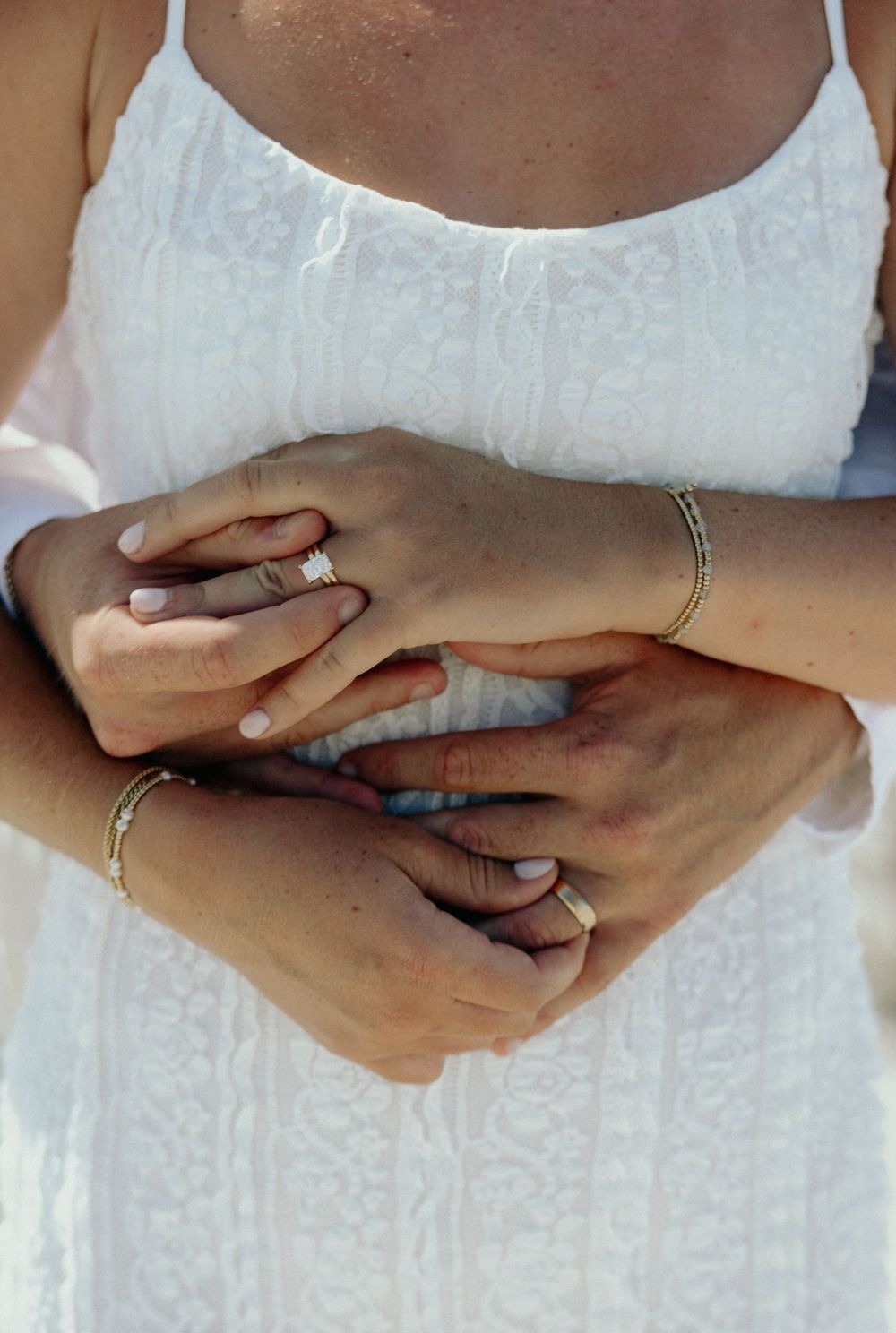 Close-up of couple holding hands showing wedding rings and bracelets against white lace dress.