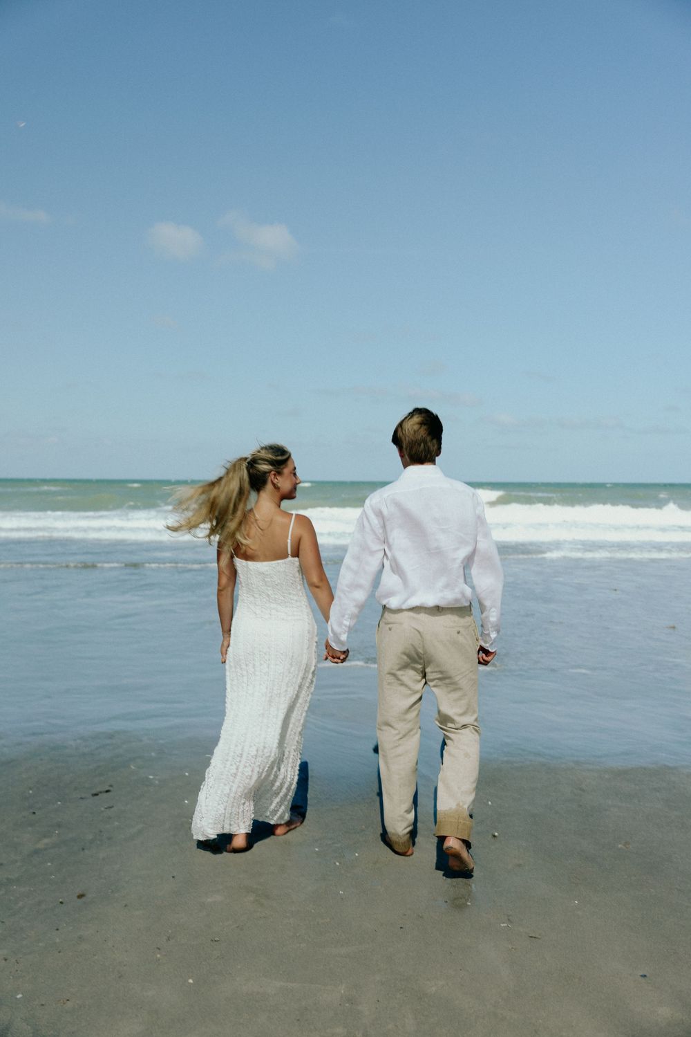 Couple in white outfits holding hands walking toward ocean waves on a sunny beach.