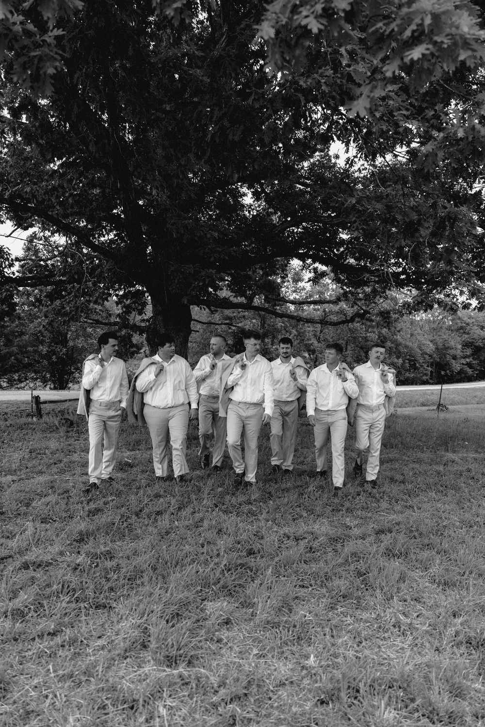 Black and white photo of groomsmen walking together in light suits under a large tree outdoors.