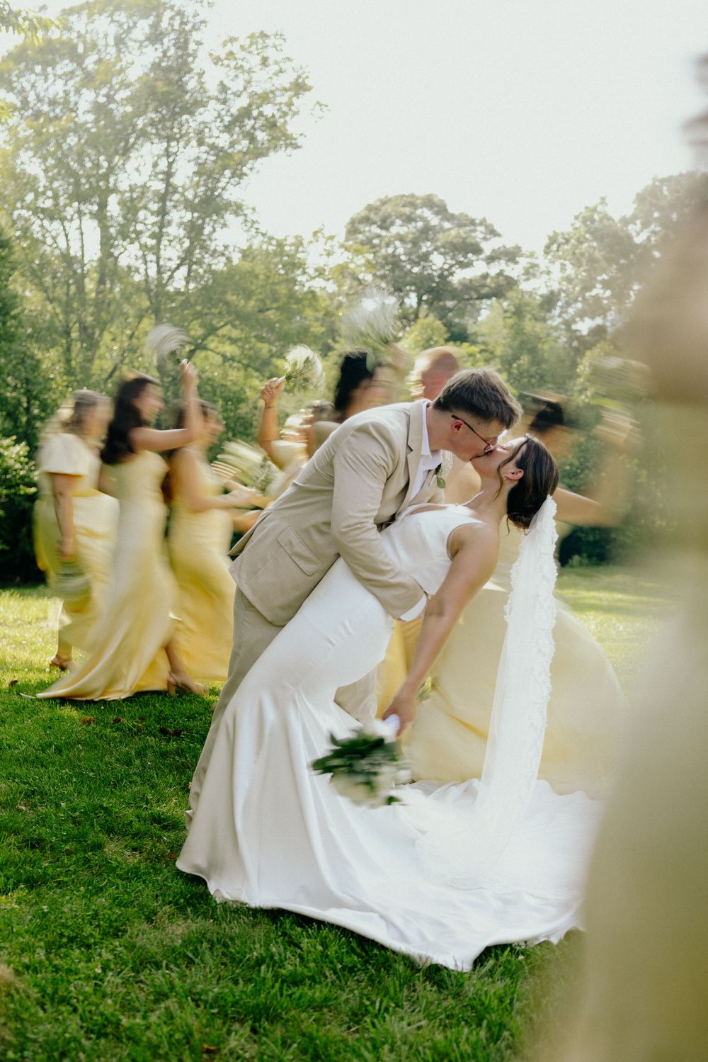 Groom dipping bride for romantic kiss surrounded by cheering wedding party in sunny outdoor setting.
