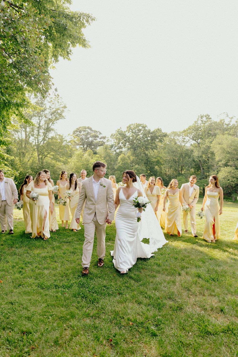 Bride and groom walking hand in hand with wedding party in yellow dresses on lush green lawn.