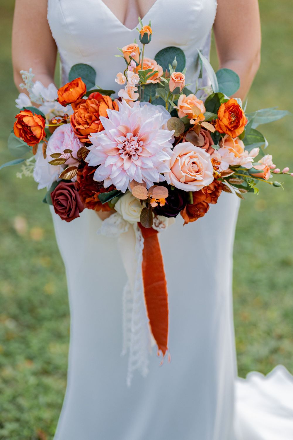Bridal bouquet featuring orange roses, pink dahlias and greenery against white wedding dress.