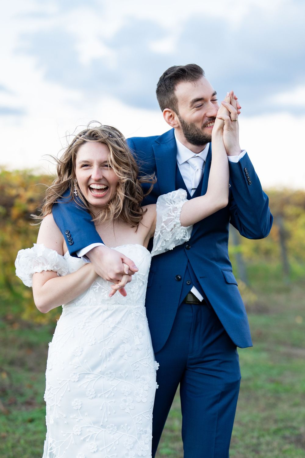 Joyful couple laughing and dancing together outdoors on their wedding day, wearing blue suit and white lace dress.