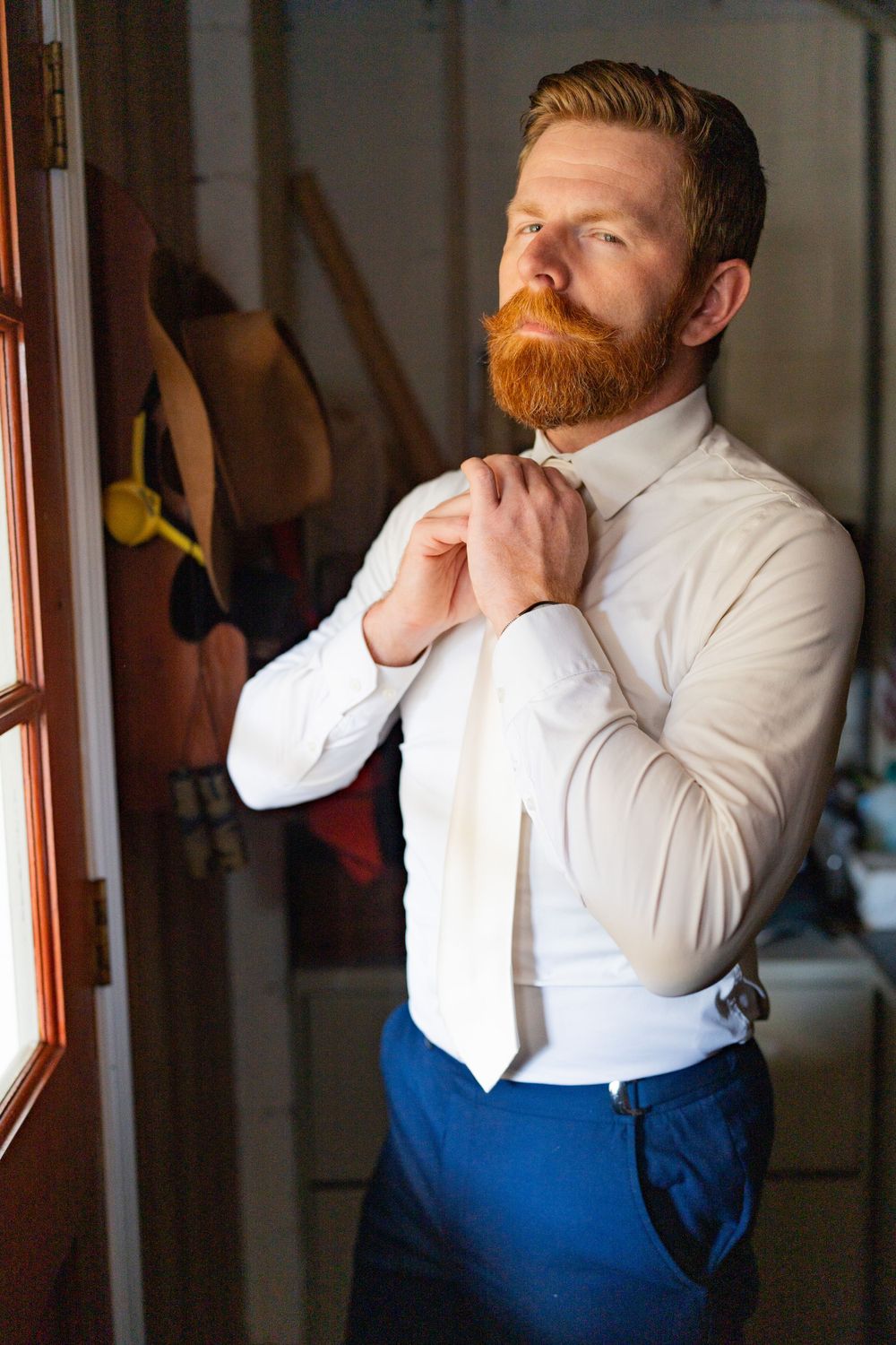 Groom adjusting tie while wearing white shirt and blue pants by window.