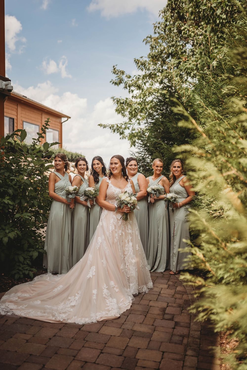 A bridal party poses outdoors in sage green bridesmaid dresses alongside a bride wearing a flowing blush lace gown.