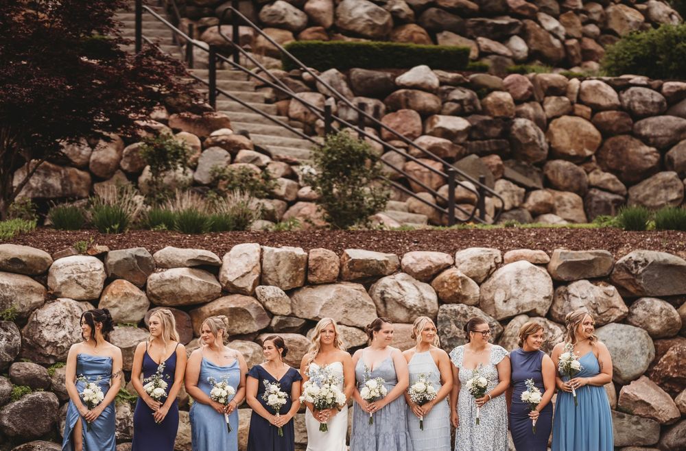 A bridal party wearing various shades of blue dresses poses in front of a rustic stone wall outdoors.