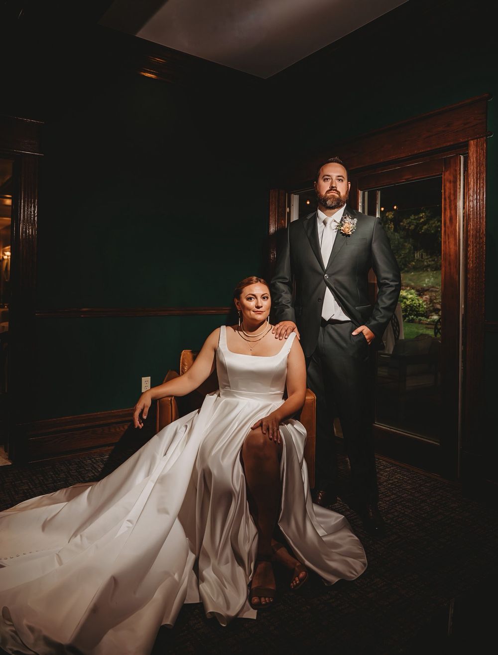 A couple in formal wedding attire poses in a dimly lit room with dark wood paneling and elegant decor.
