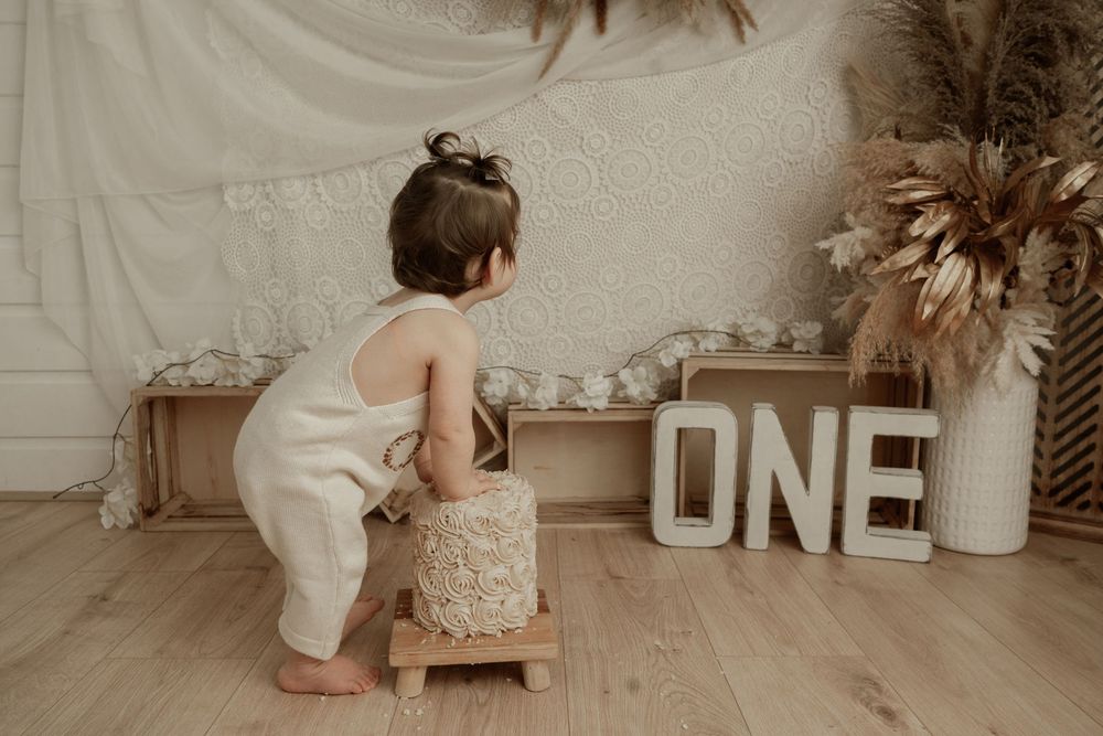 A baby in white romper crawls on hardwood floor near large decorative letters and toys in a neutral-toned room.