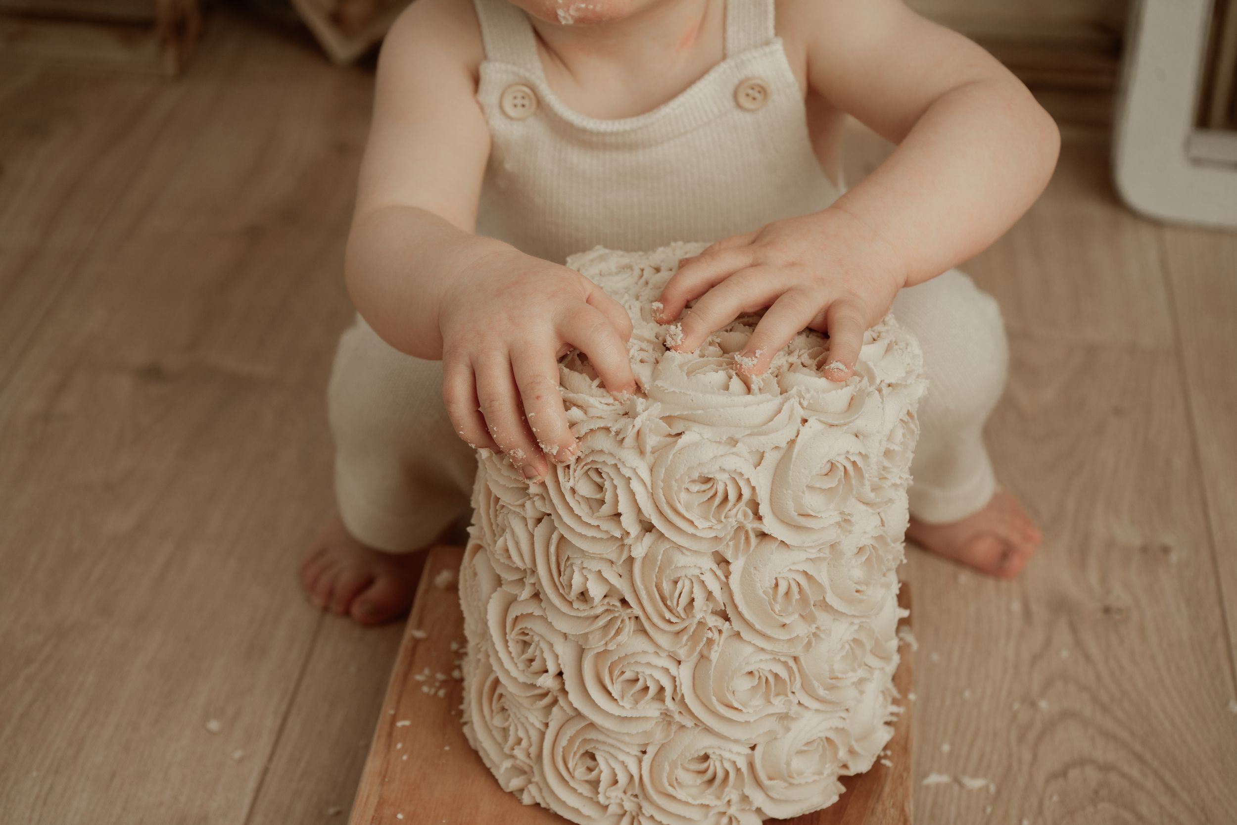 Small hands reach for a delicate white frosted cake with rosette swirl details on a wooden floor.