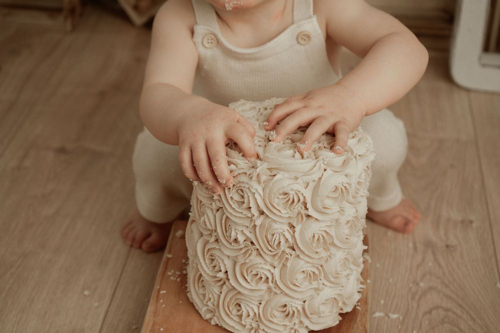 Small hands reach for a delicate white frosted cake with rosette swirl details on a wooden floor.