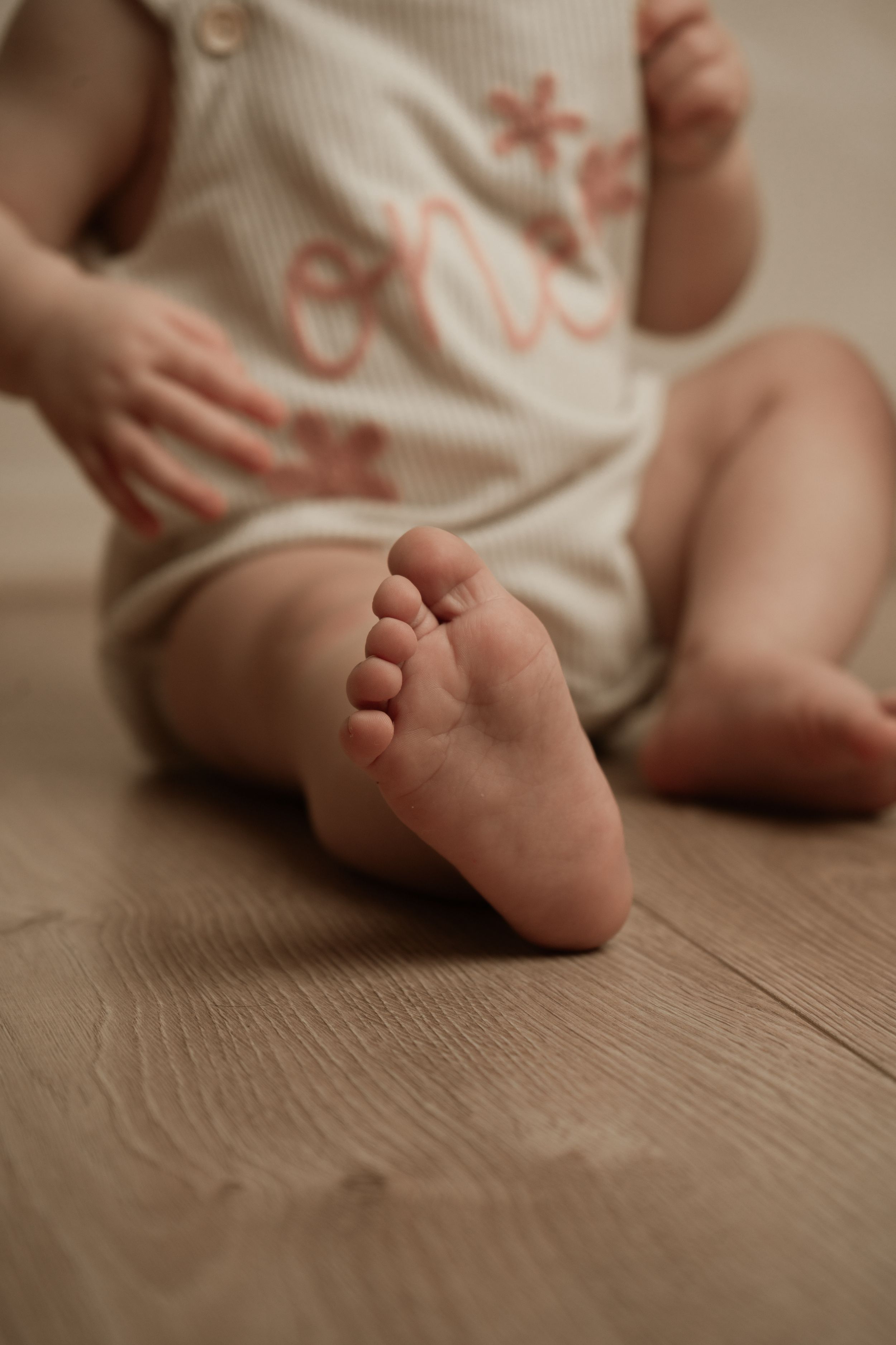 Close-up of tiny baby's foot on wooden floor with cream-coloured onesie visible in soft, warm lighting.