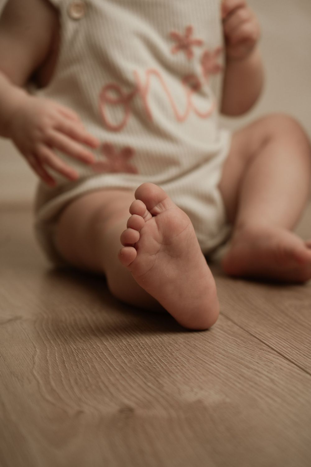 Close-up of tiny baby's foot on wooden floor with cream-coloured onesie visible in soft, warm lighting.
