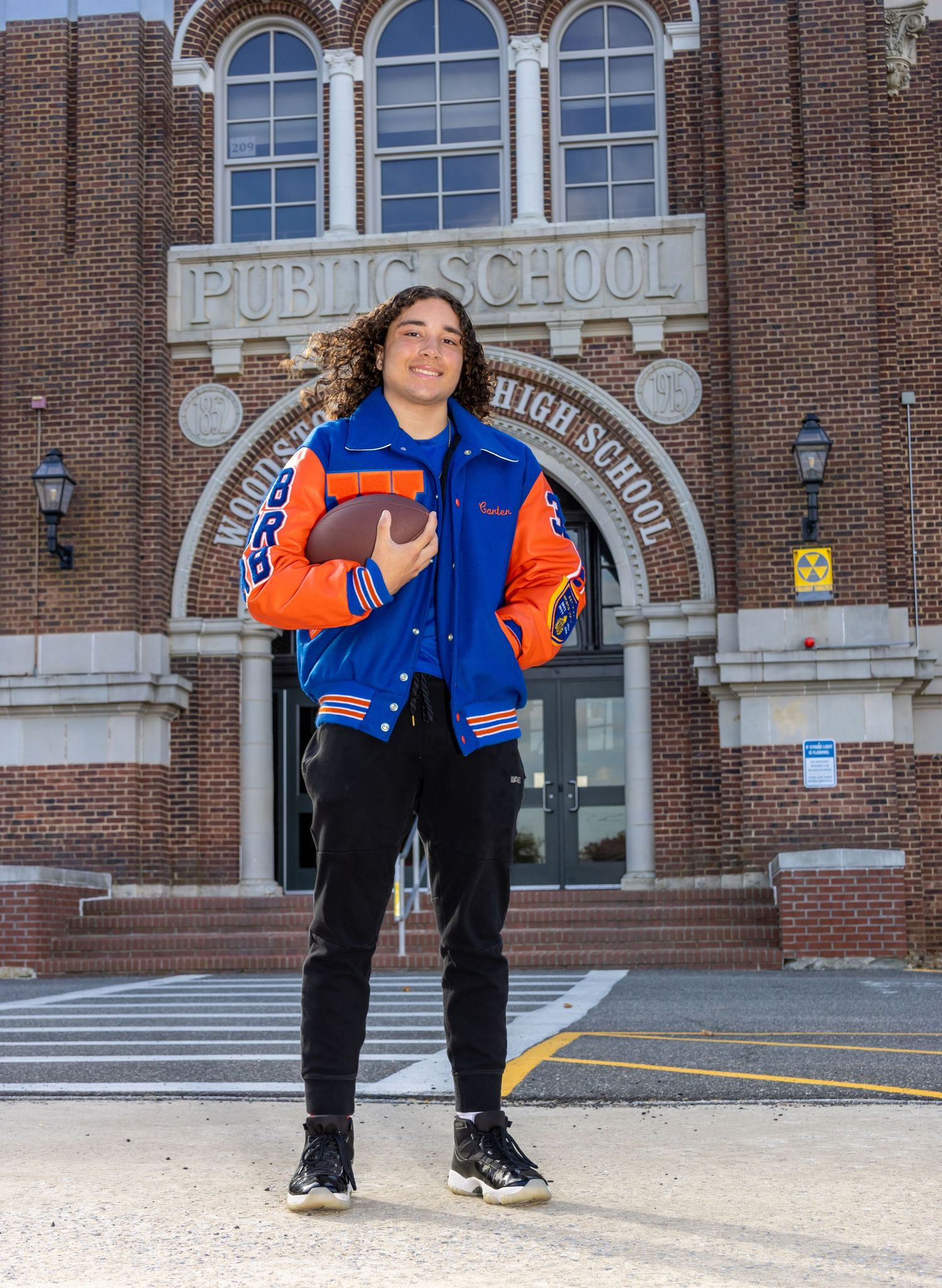 Senior boy holding a football in his letterman jacket out front of his school. Salem and Gloucester County Senior Portraits